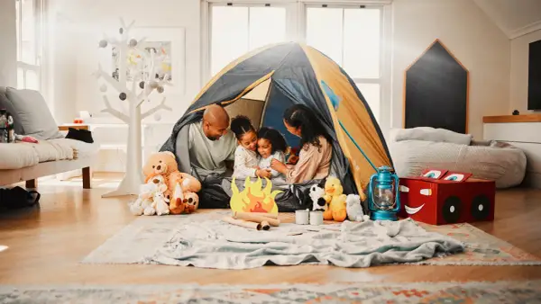 Familie verbringt **gemeinsame Zeit** beim Zelten im Wohnzimmer. Kinder kuscheln mit Teddybären, während Eltern lachen.