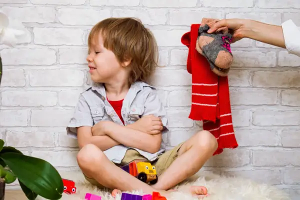 Ein Junge sitzt mit verschränkten Armen und einem trotzigen Gesichtsausdruck da. Über ihm hält eine Hand einen roten Schal und ein Paar Socken. Der Junge will sich nicht anziehen.